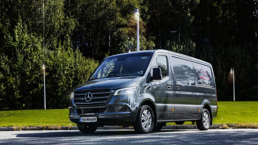 Fronten av en mørkegrå budbil med Road Runners logoen på siden. Masse trær og lys i bakgrunnen.